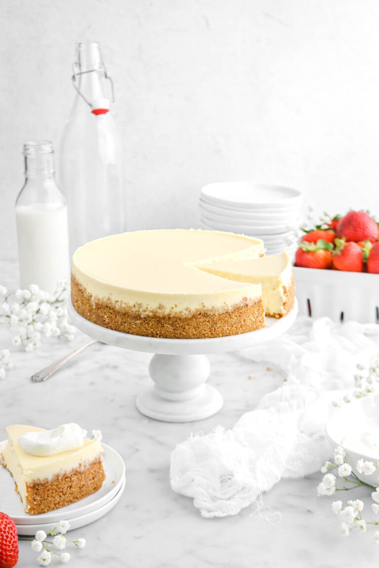 cheesecake with slice on marble cake stand, flowers and white napkin beside, and second slice of cheesecake on two stacked white plates