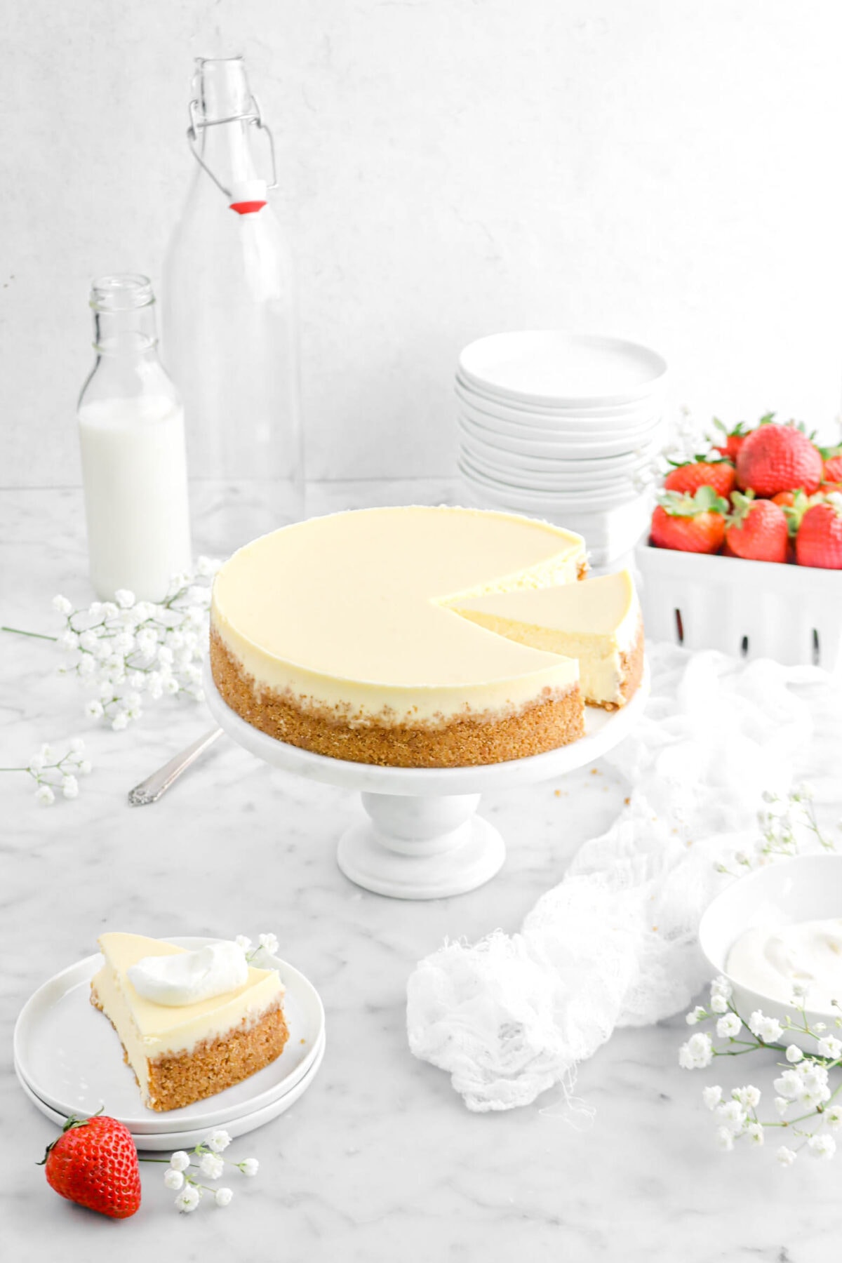 angled shot of cheesecake with slice cut on marble cake stand with flowers, strawberries, and stack of plates beside, a second slice of cheesecake beside on two stacked plates