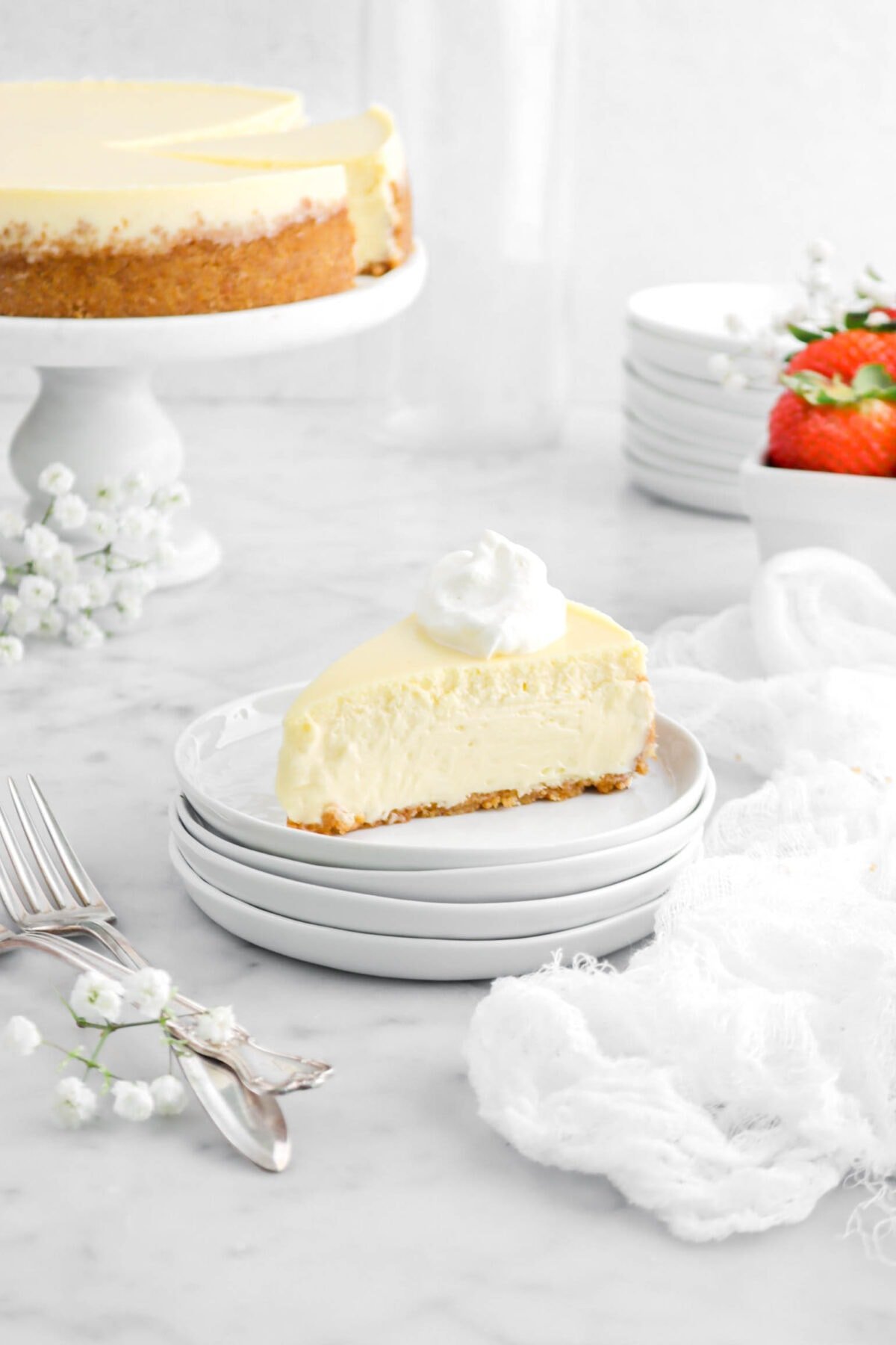 slice of cheesecake on four stacked plates with dollop of whipped cream on top, forks and white napkin beside, cheesecake on cake stand and strawberries behind