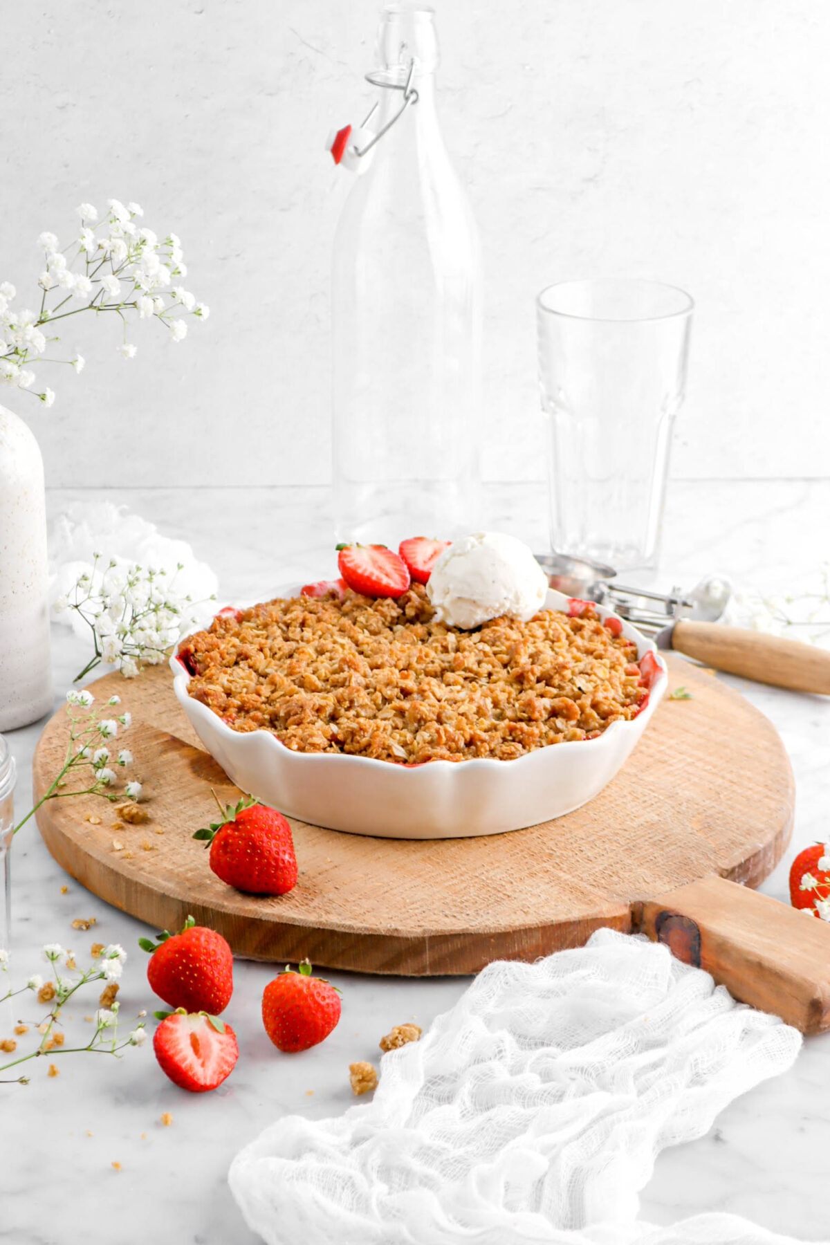 front shot of strawberry rhubarb crisp on wood board with white cheese cloth on marble surface with strawberries and flowers around