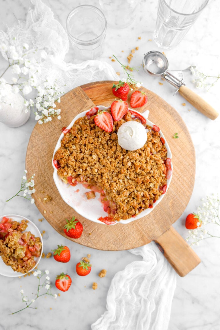 strawberry rhubarb crisp on round wood board with vanilla ice cream on top and fresh strawberries around, an ice cream scoop beside, a white cheese cloth, and plate of crisp beside