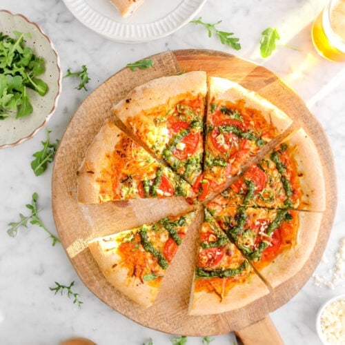 sliced pizza on wood board with fresh arugula around, slice of pizza on white plate above with glass of beer on marble surface