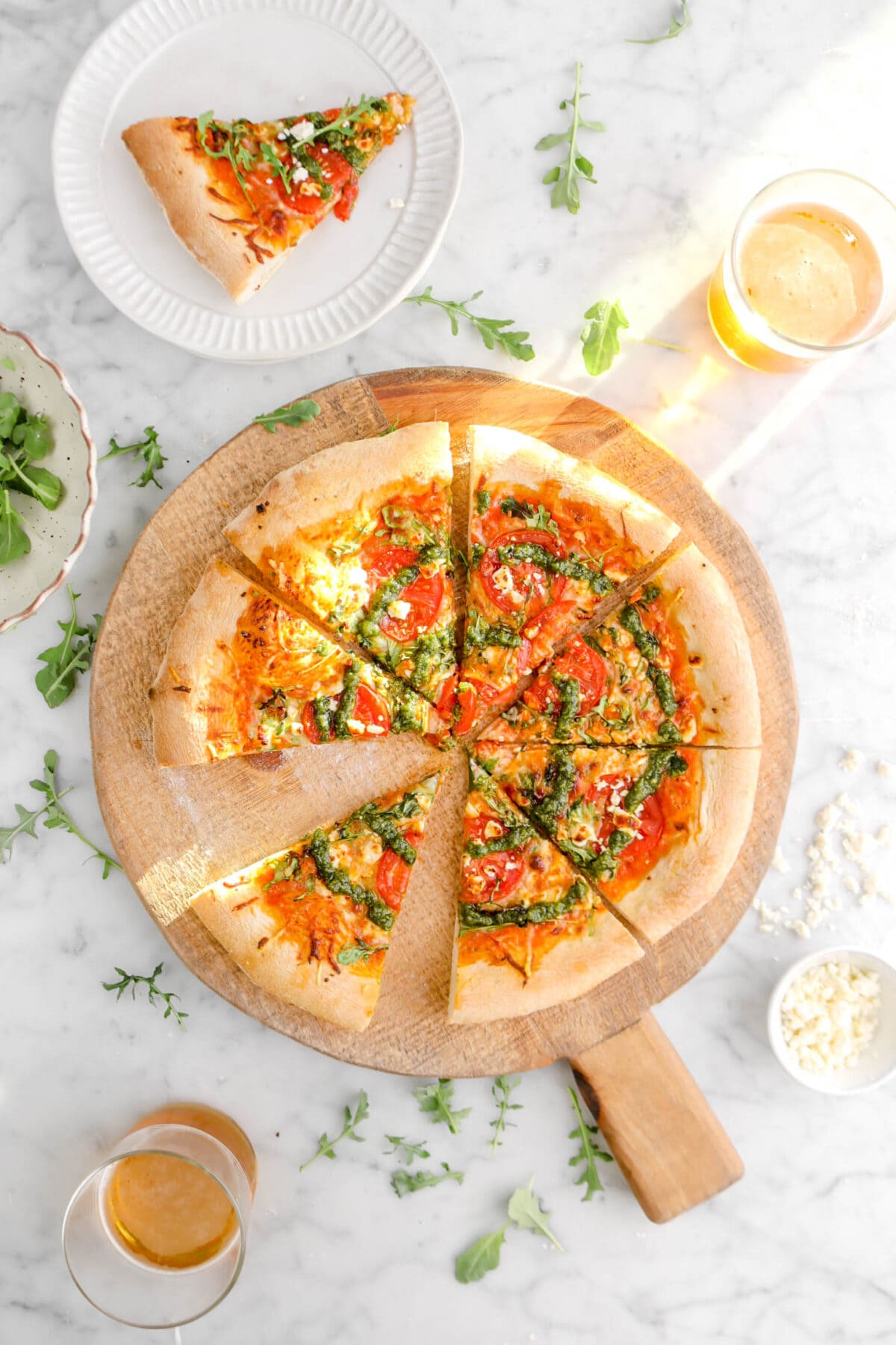pizza on round wood board with slice on stack of plates beside, with arugula beisde, two glasses of beer, and bowl of crumbles feta