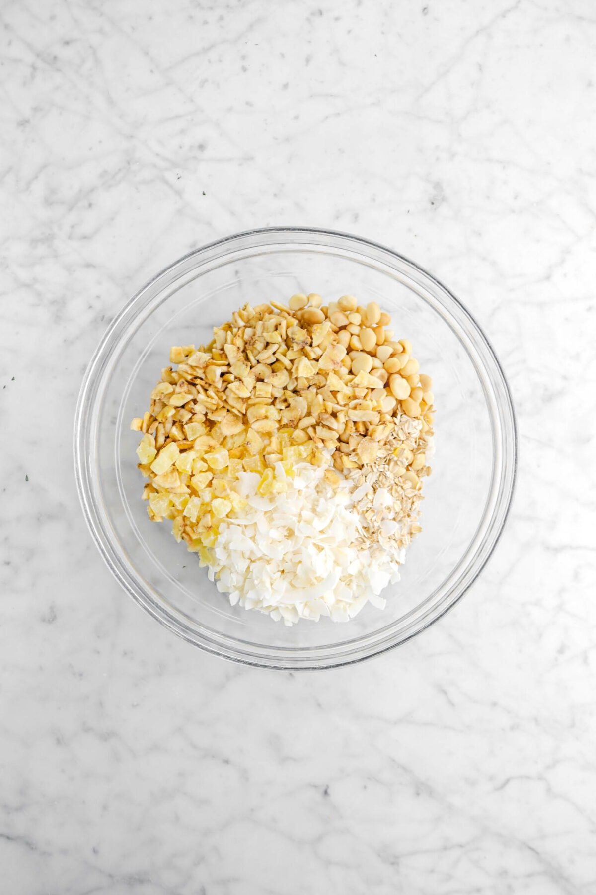 oats, coconut flakes, dried pineapple, banana chips, and macadamia nuts in glass bowl on marble surface