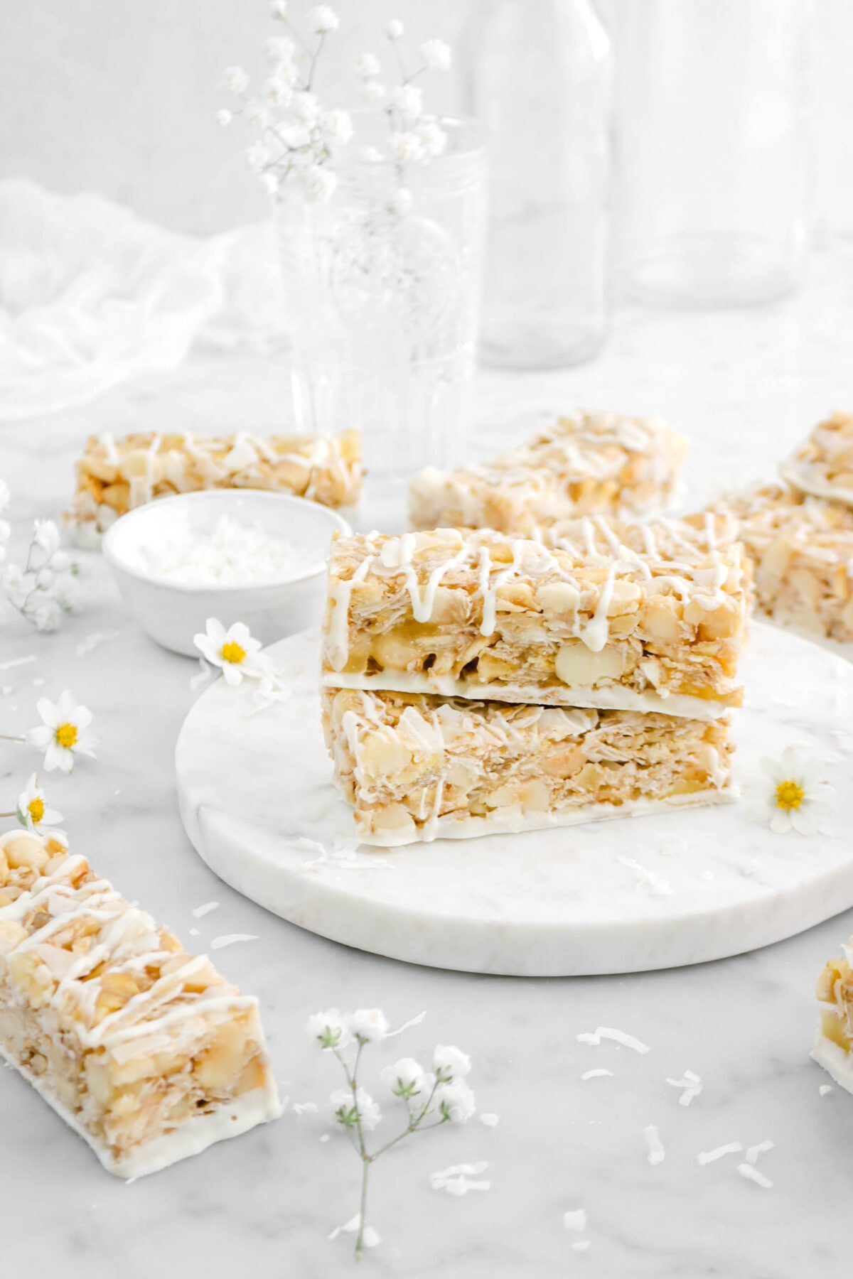 angled shot of two stacked granola bars on marble plate with more granola bars around, flowers, and shredded coconut