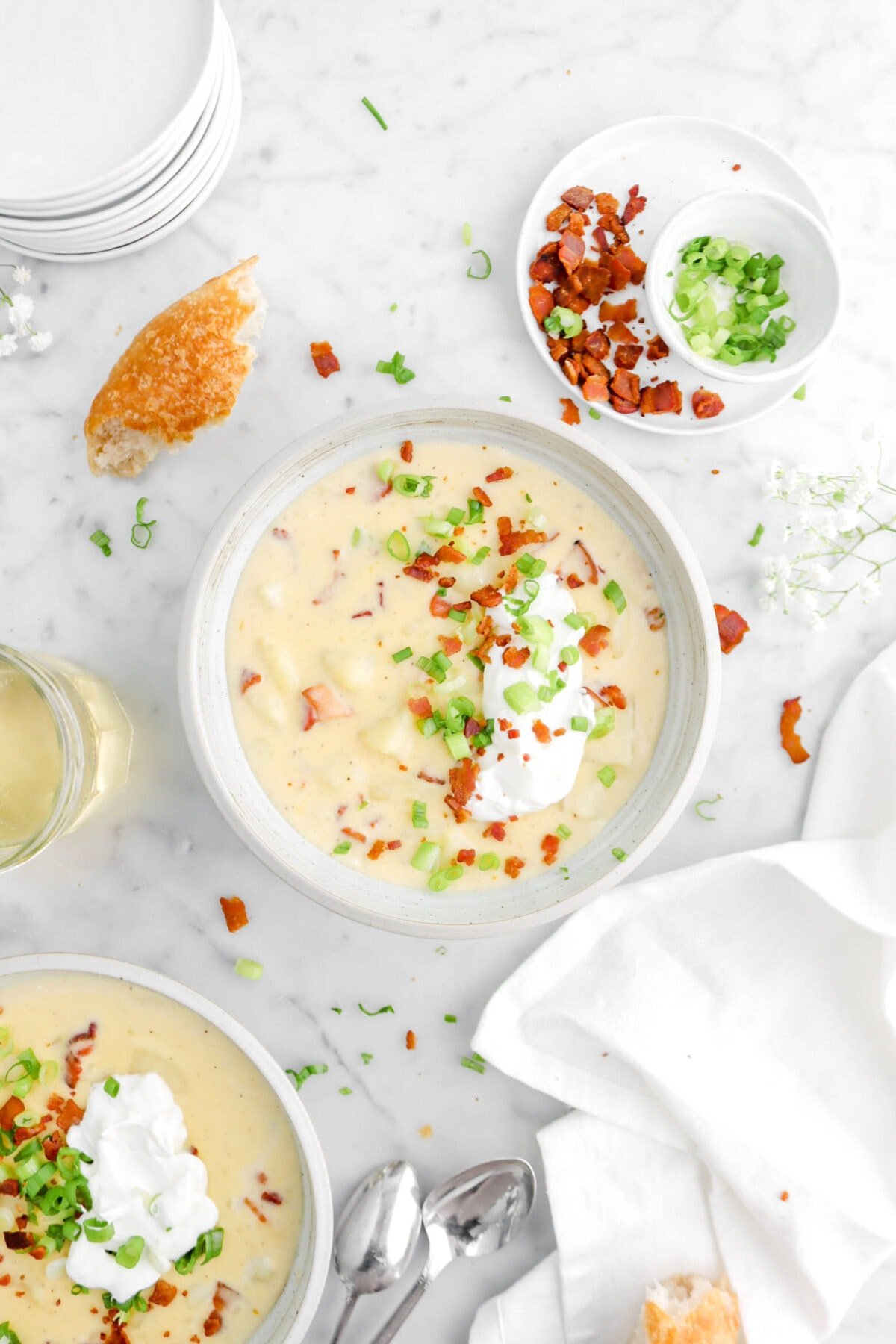 bowl of baked potato soup on marble surface with chopped bacon, chopped green onion, and sour cream on top with white napkin, chunk of bread, and another bowl beside
