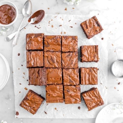 sixteen brownies on parchment paper with bowl of melted chocolate and two spoons beside, glass of milk, measuring cup of salt, and plates on marble surface