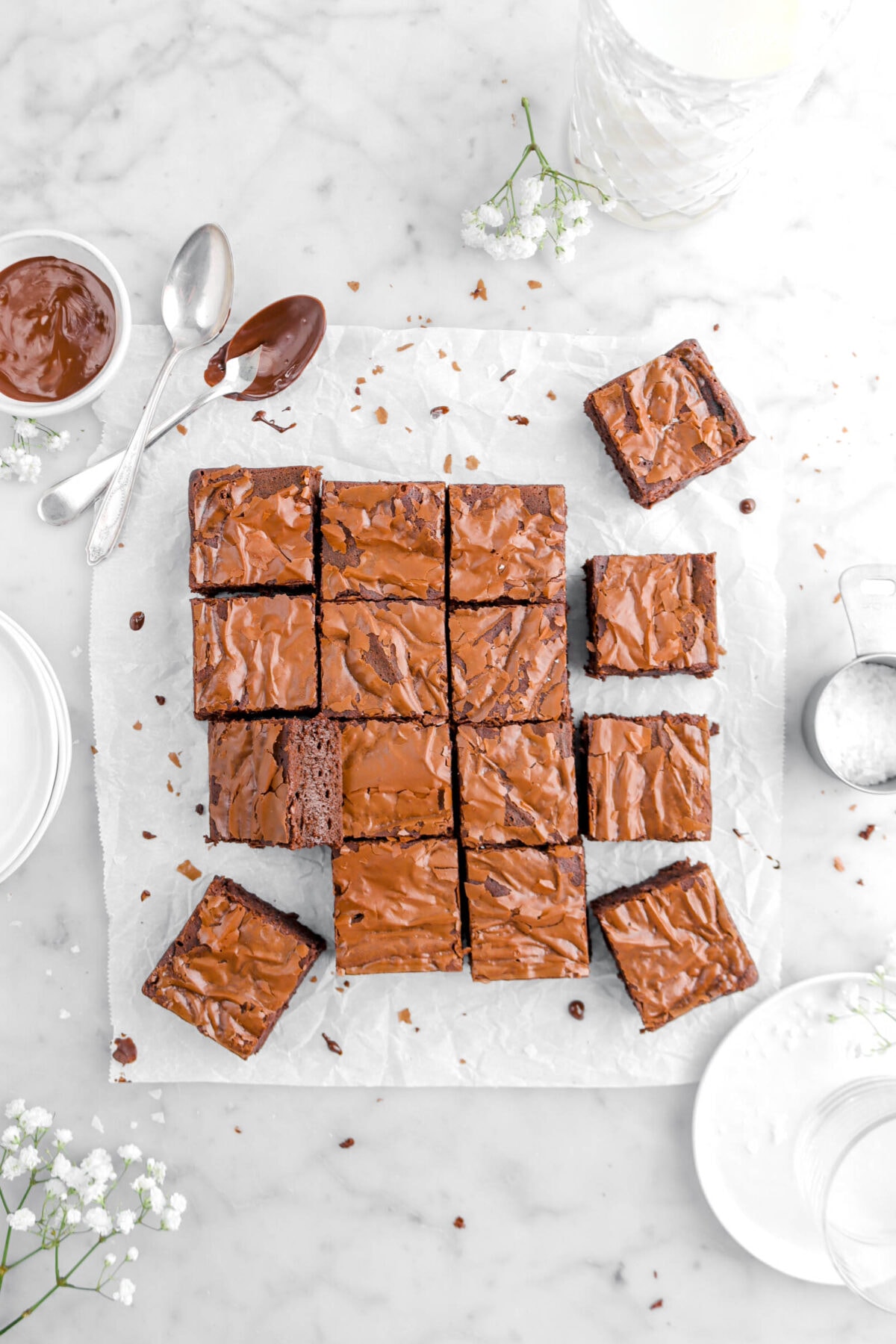 sixteen brownies on parchment paper with bowl of melted chocolate and two spoons beside, glass of milk, measuring cup of salt, and plates on marble surface