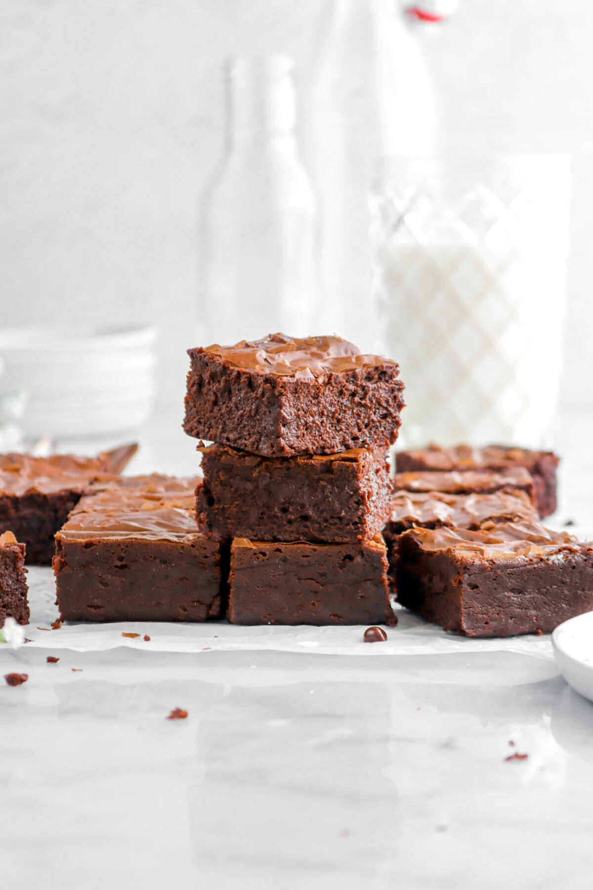 three stacked brownies on parchment paper with more brownies and glass of milk behind