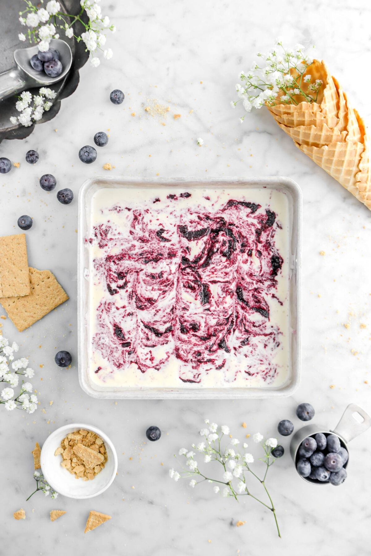square pan of blueberry cheesecake ice cream on marble surface with graham crackers around, blueberries, and white flowers