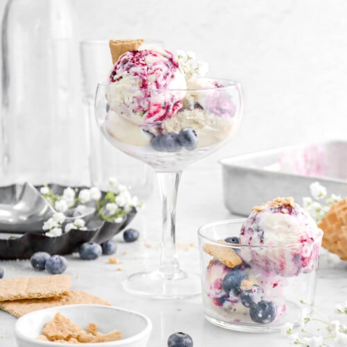 blueberry cheesecake ice cream in coupe glass with smaller glass beside with graham crackers and blueberries on top, blueberries, graham crackers, and white flowers around with two empty glasses behind