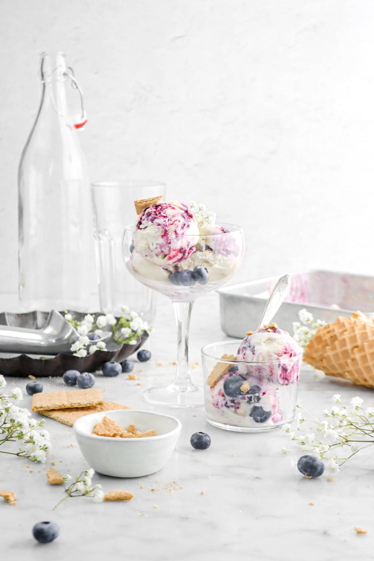 pulled back shot of ice cream in coupe glass with smaller glass beside and spoon inside, flowers, blueberries, and graham crackers on marble surface with pie pan and cake pan behind