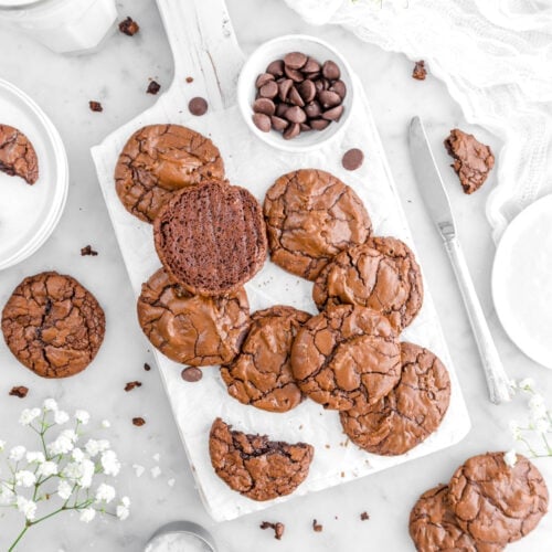 overhead shot of brownie cookies on white wood board with bowl of chocolate chips beside, cookies around, knife beside, glass of milk, flowers, and cookie crumbs around on marble surface