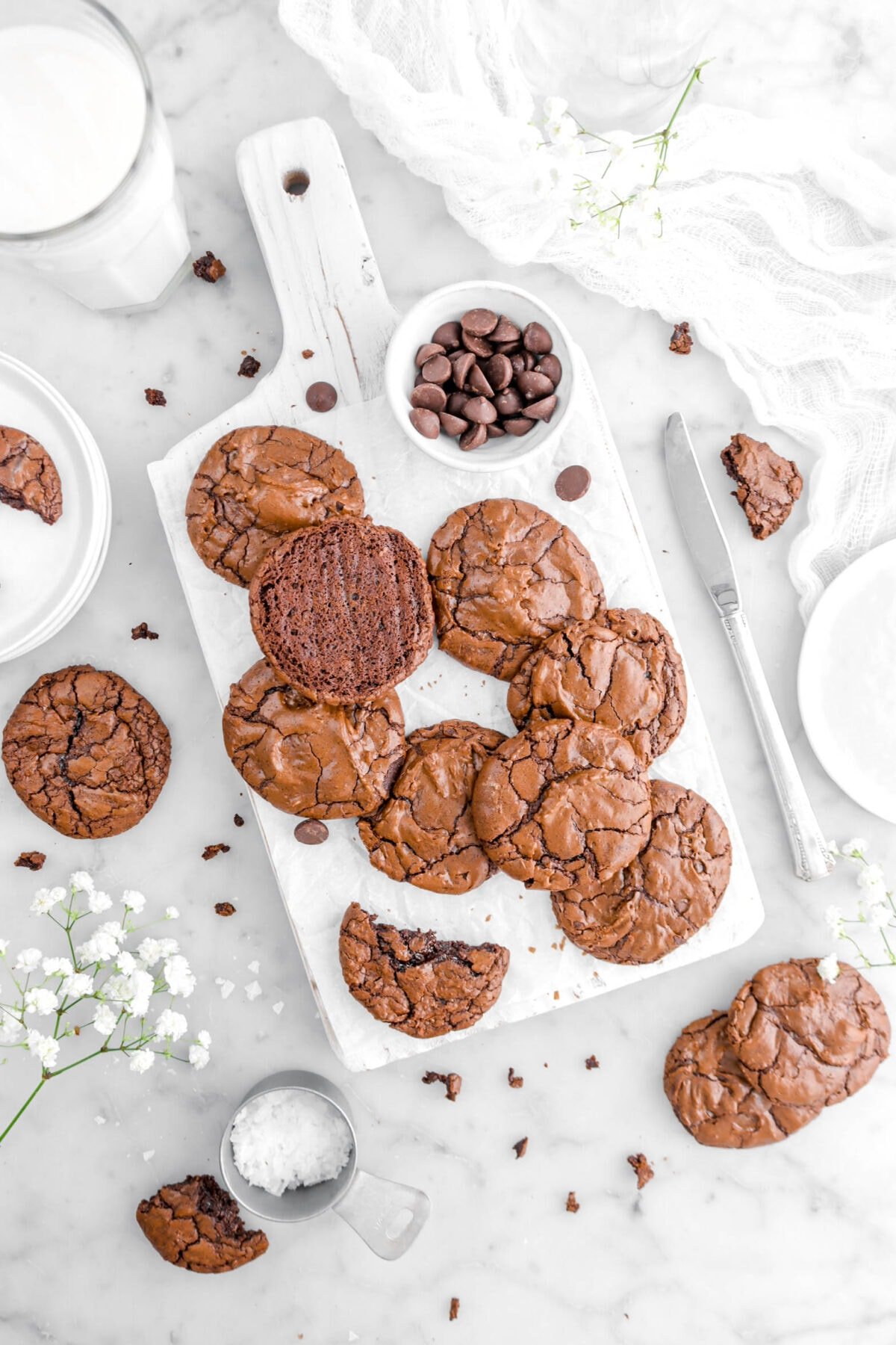 overhead shot of brownie cookies on white wood board with bowl of chocolate chips beside, cookies around, knife beside, glass of milk, flowers, and cookie crumbs around on marble surface