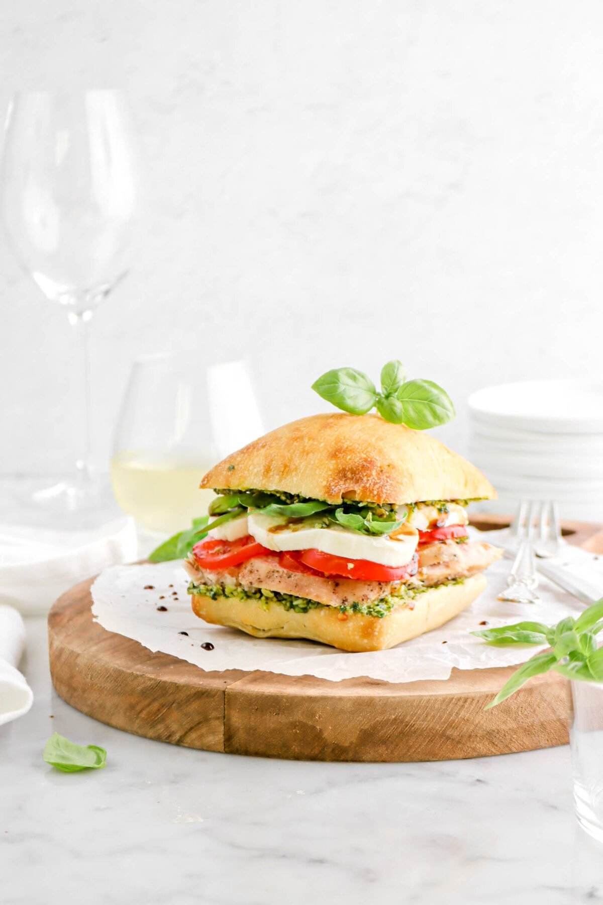 chicken caprese sandwich on parchment paper on wood board with sprig of basil on top, two wine glasses behind, and a stack of white plates behind
