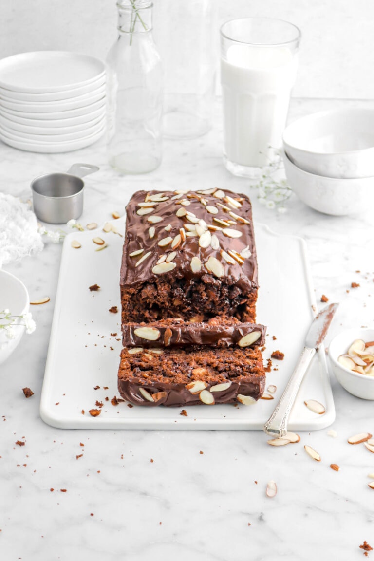 angled shot of chocolate loaf cake on white tray with melted chocolate and sliced almonds on top, a knife beside, flowers, two coffee mugs, and glass of milk behind