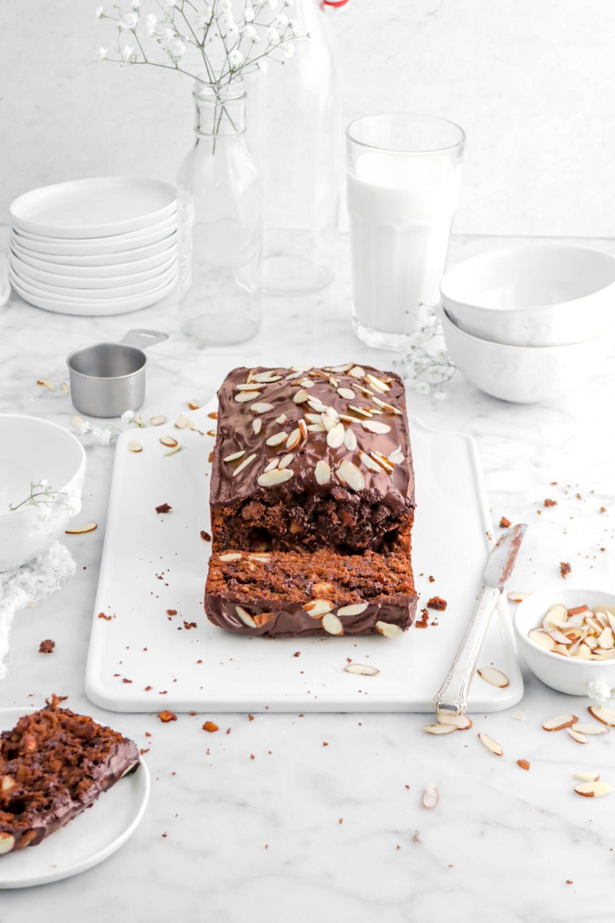 pulled back angled shot of chocolate loaf on white tray with slice laying on front, plate with slice beisde, bowl of sliced almonds beside, glass of milk and stack of plates behind