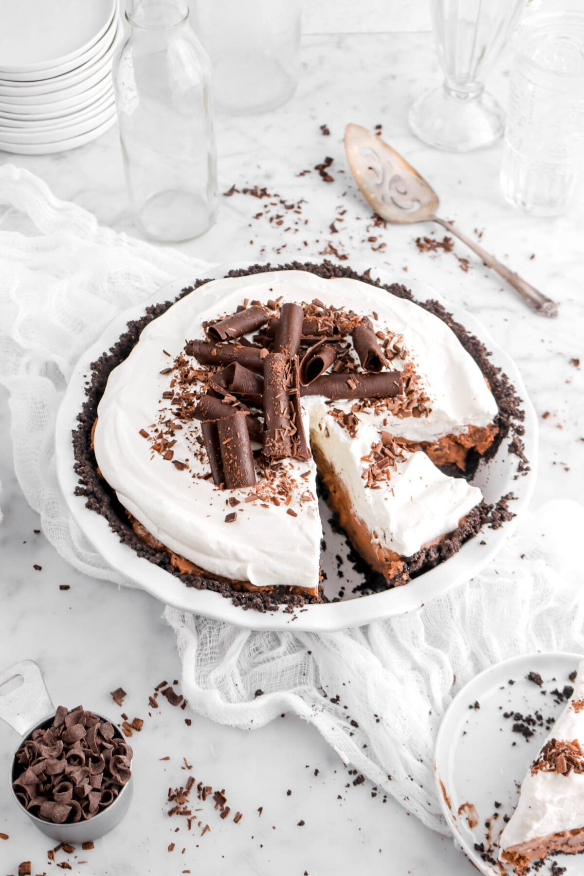 angled shot of mousse pie with a slice still inside pie plate with large chocolate curls, chocolate shavings, and slice of pie beside on white cheesecloth