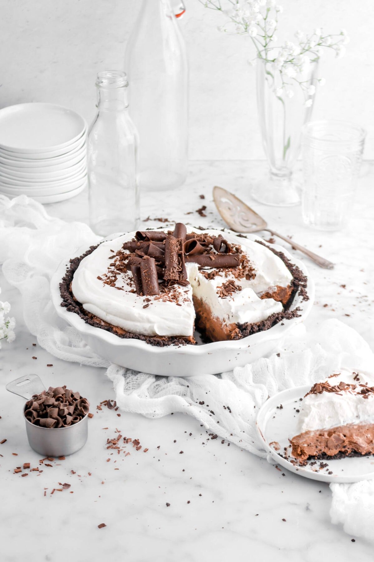 chocolate mousse pie on white cheese cloth with whipped cream and large chocolate curls on top, with slice on white plate in front, a measuring cup of chocolate curls, stack of plates beside, and empty glasses