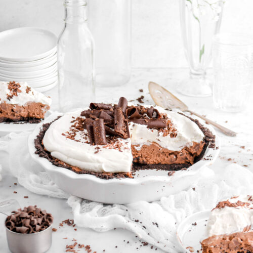 angled shot of chocolate mousse pie with two slices in front and behind on white plates, chocolate shavings around, a white cheese cloth, and empty glasses behind