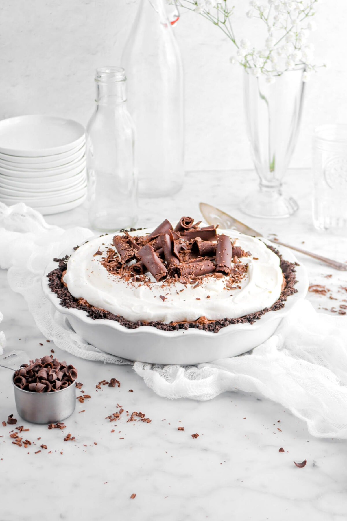 pie on white cheese cloth with large chocolate curls and whipped cream on top, empty glasses, and more chocolate curls around