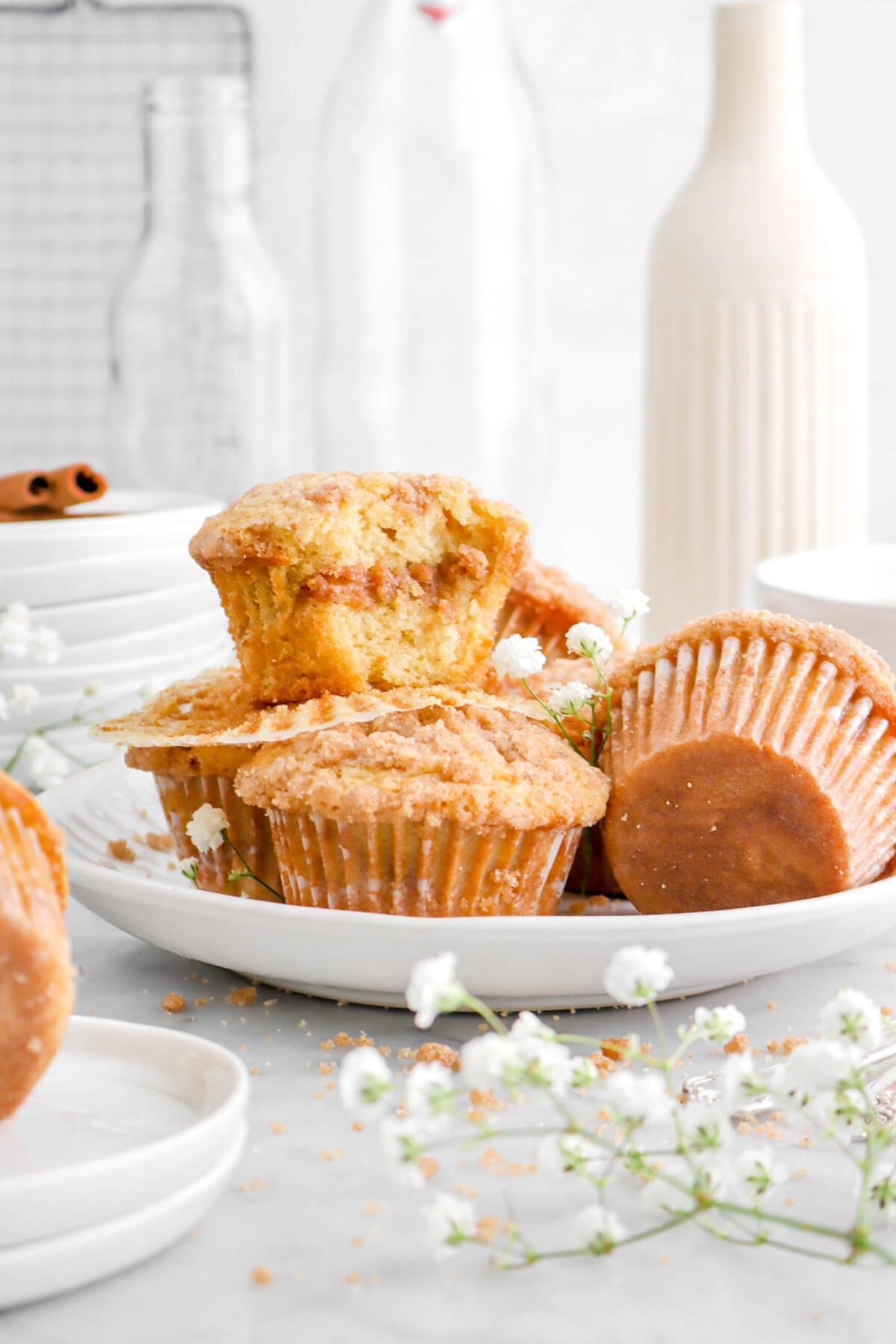 coffee cake muffins on white plate with bite missing from one, white flowers, vase, and empty glasses behind