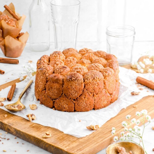 cinnamon sugar walnut pull apart bread on crumbled parchment paper on a wood board with walnuts, cinnamon sticks, a gold spoon and bowl beside, a stack of muffins, an empty glass, and flowers behind