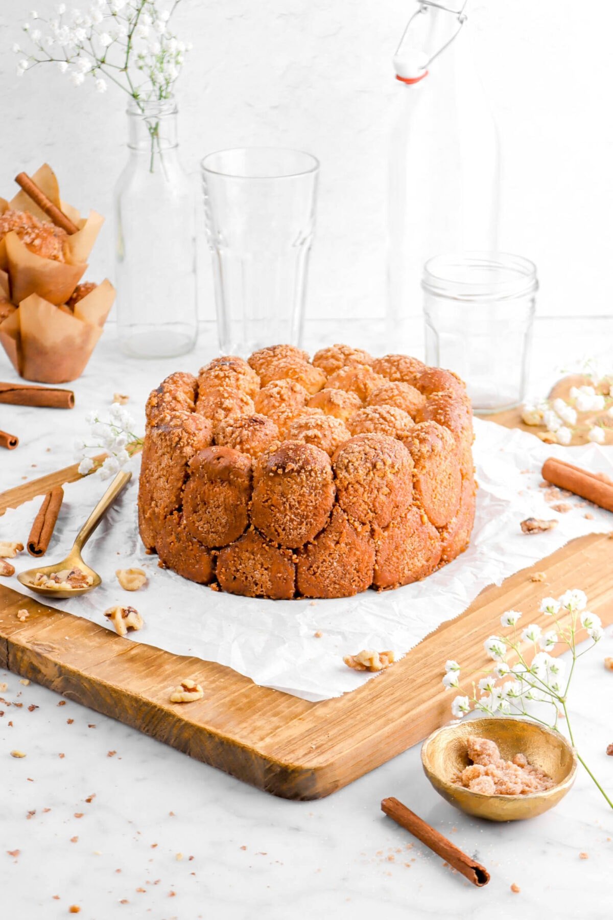 cinnamon sugar walnut pull apart bread on crumbled parchment paper on a wood board with walnuts, cinnamon sticks, a gold spoon and bowl beside, a stack of muffins, an empty glass, and flowers behind