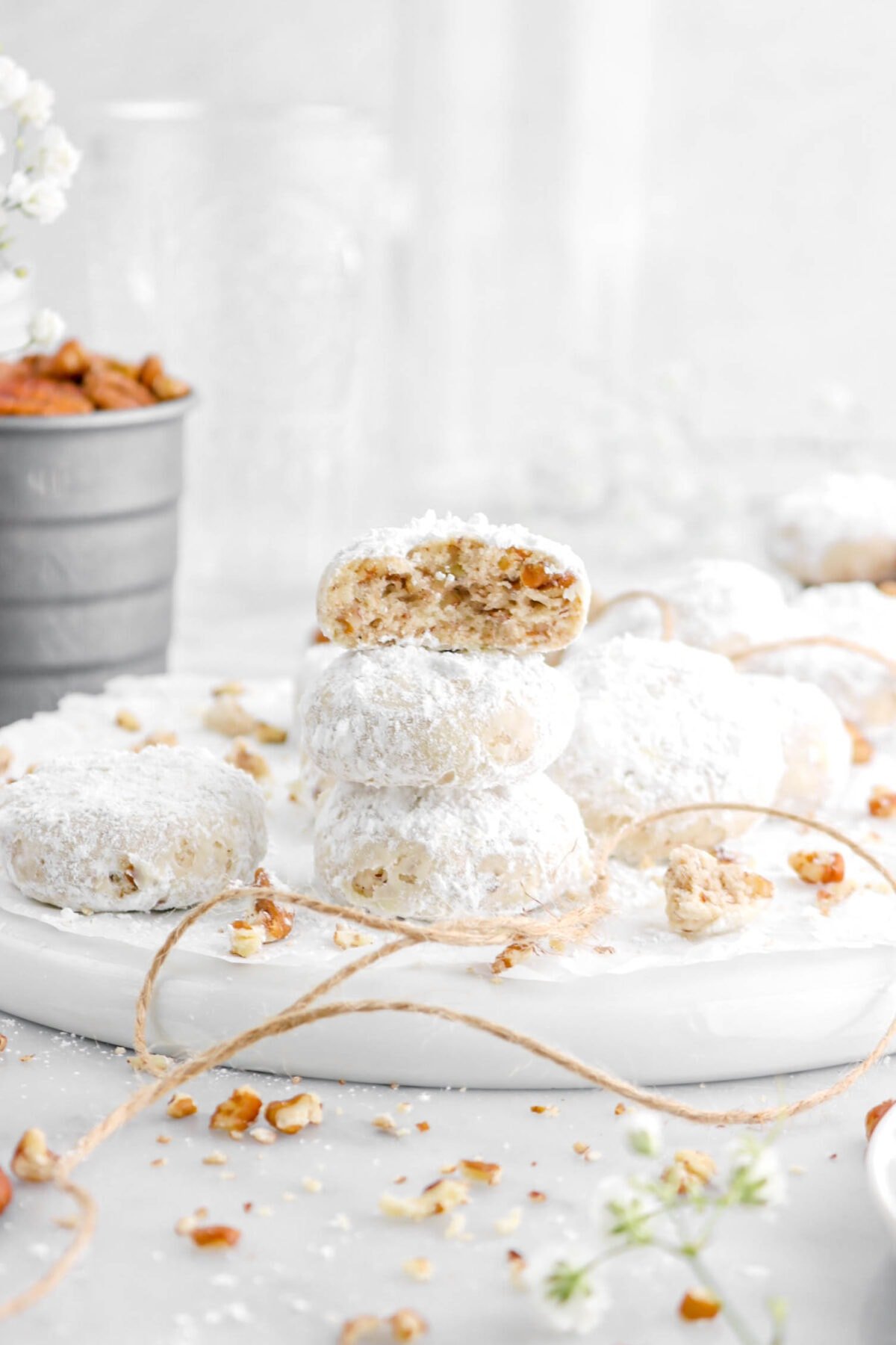 three stacked danish wedding cookies on upside down white plate with white flowers behind and measuring cup of pecans