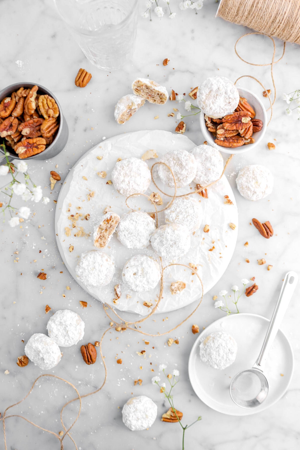 nine danish wedding cookies on white plate with one broken in half, more cookies around on marble surface with tan string