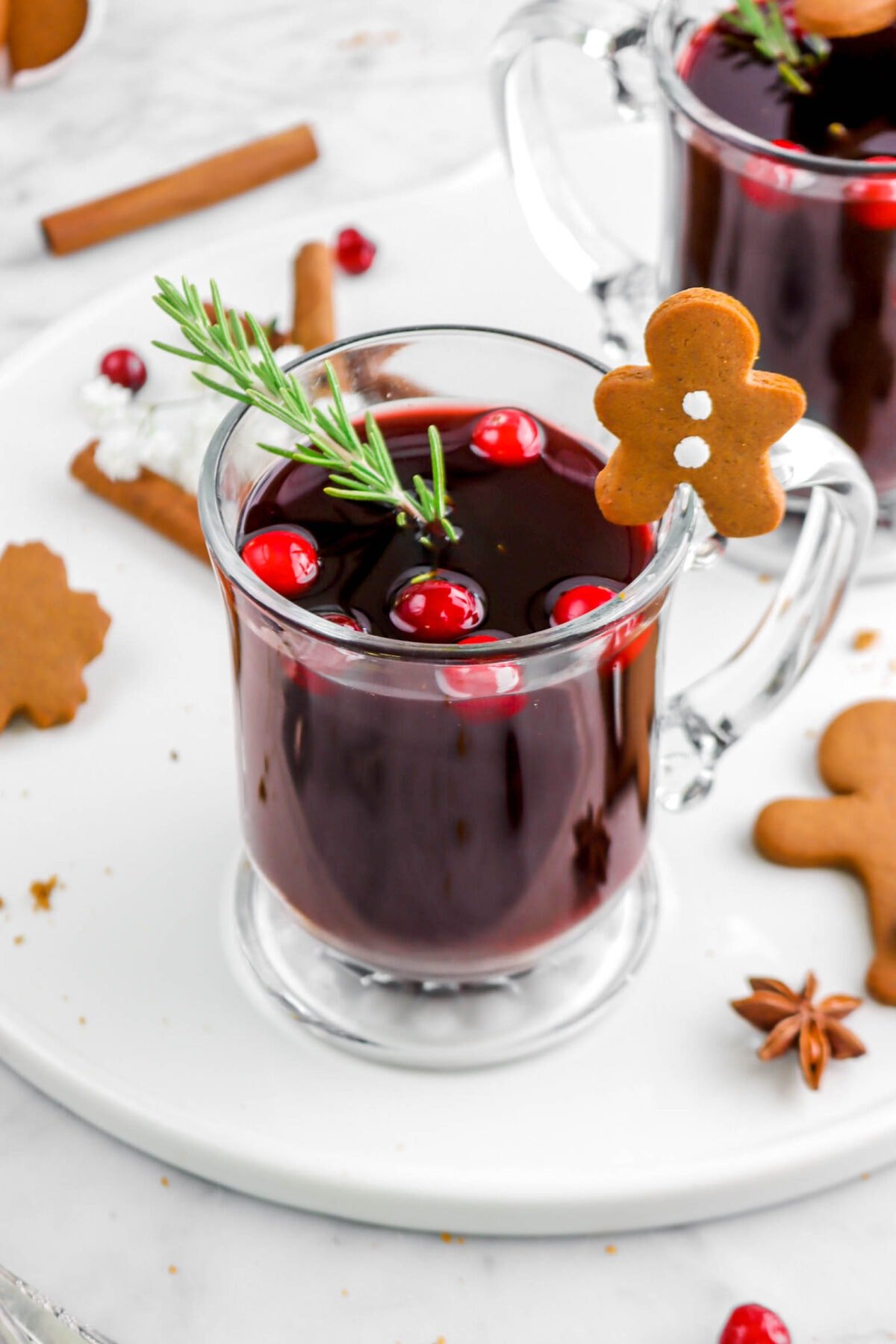 close up of mulled wine in glass mug with sprig of rosemary and six cranberries in the drink, and a gingerbread cookie on the rim