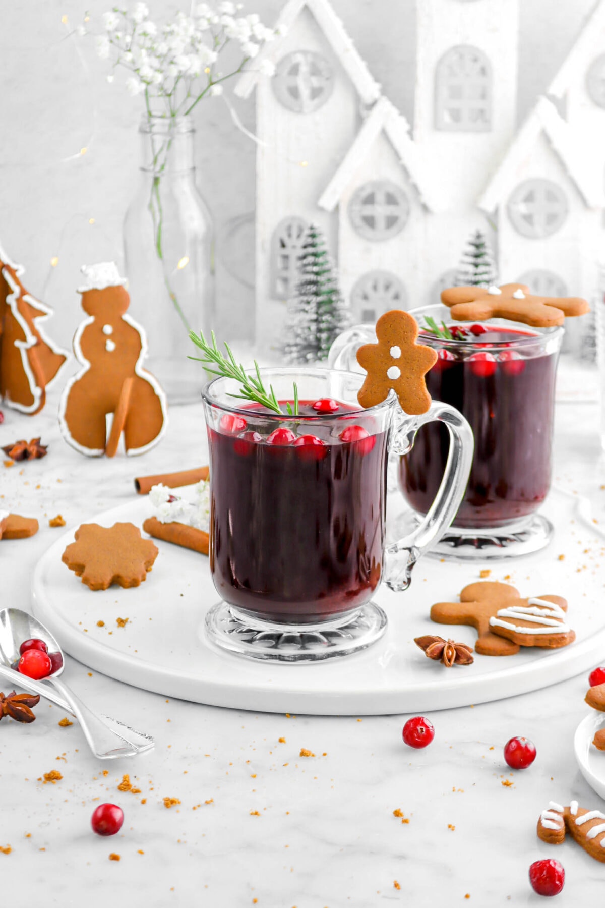 front shot of mulled wine in two glass mugs on white tray with christmas village and white flowers behind, gingerbread cookies around, whole spices, and cranberries beside