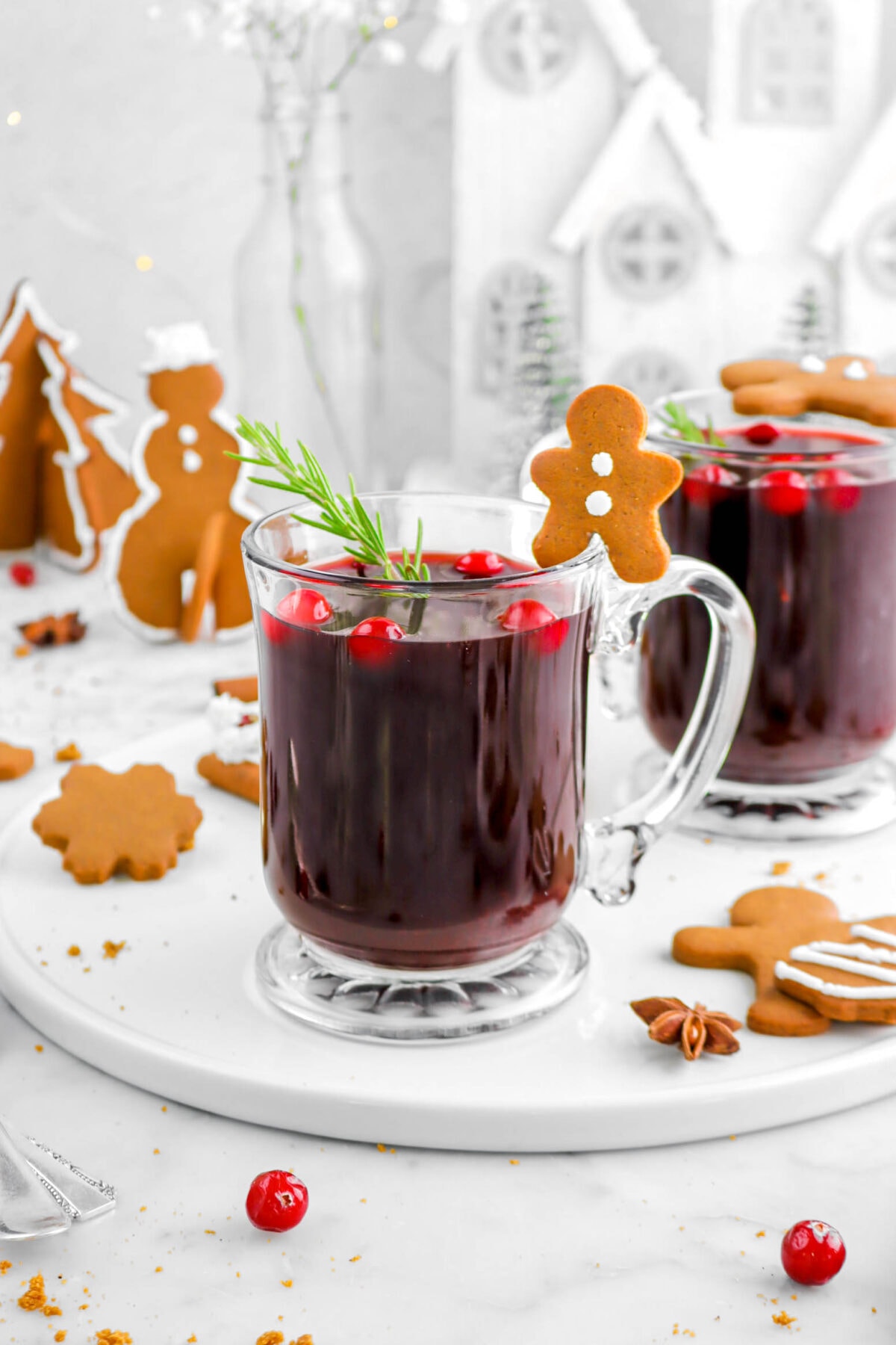 mulled wine in two glass mugs on white serving tray with cranberries and rosemary sprigs with gingerbread cookies around