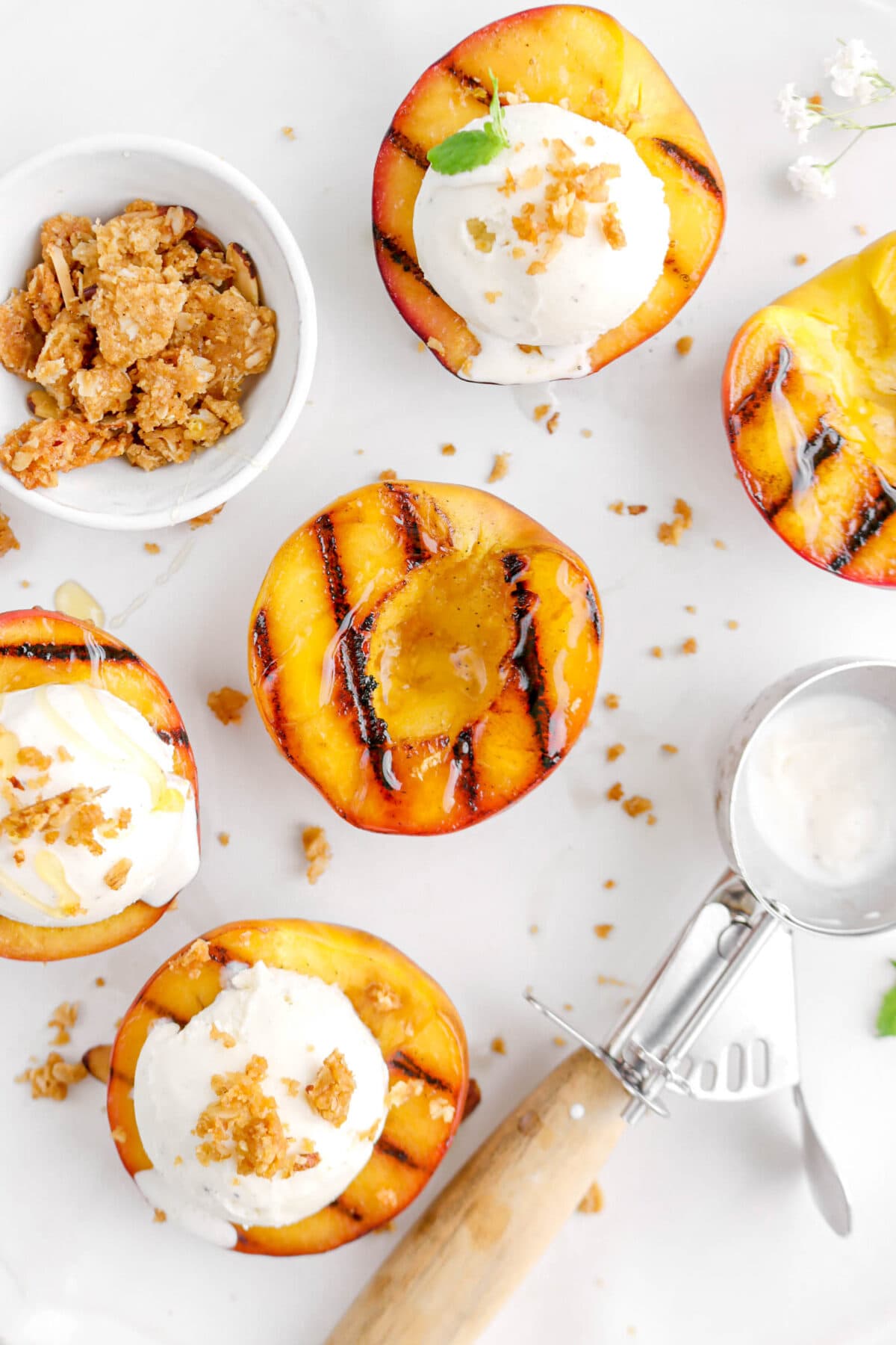 close up of five grilled peaches, three with scoops of ice cream on top, and bowl of crumble beside