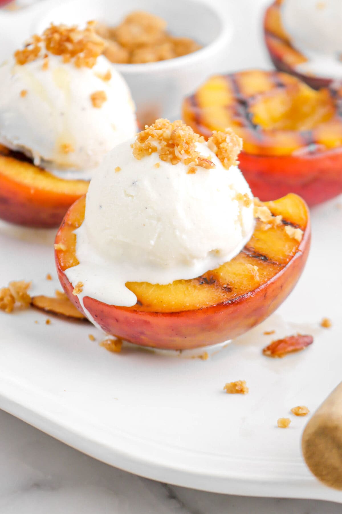 close up of grilled peach on white platter with scoop of ice cream on top and crumble