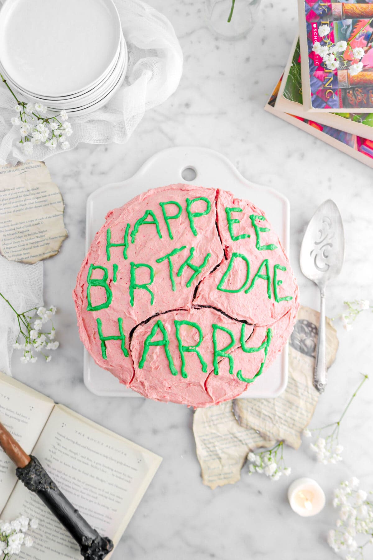overhead shot of cake reading "happy birthday harry" misspelled in green frosting with book pages and flowers around