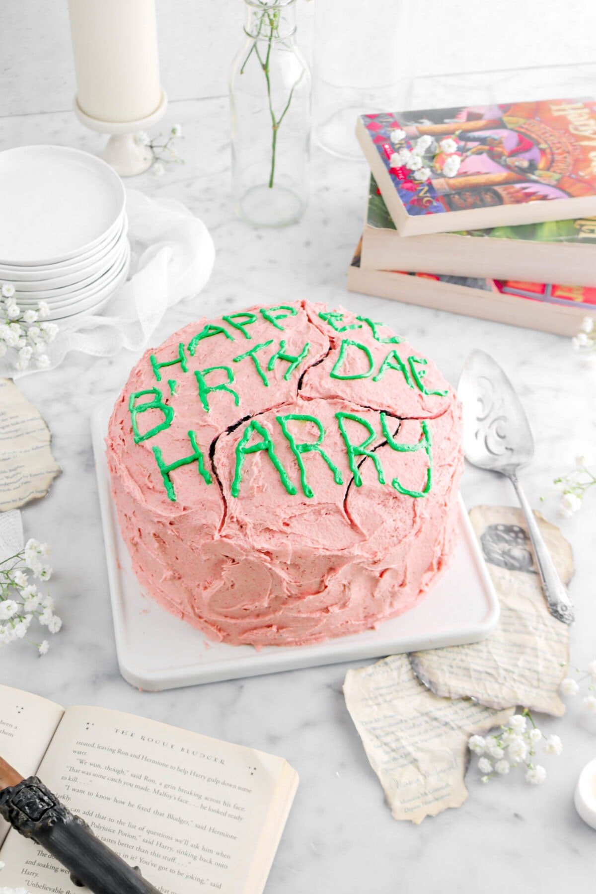 close up angled photo of cake with strawberry frosting and green piping on top, burned book pages around with white flowers and a cake knife beside