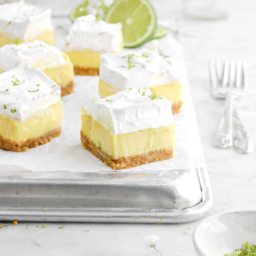 six key lime pie bars on sheet pan with sliced lime behind with white flowers, forks beside, and small white bowl of lime zest in front on marble surface