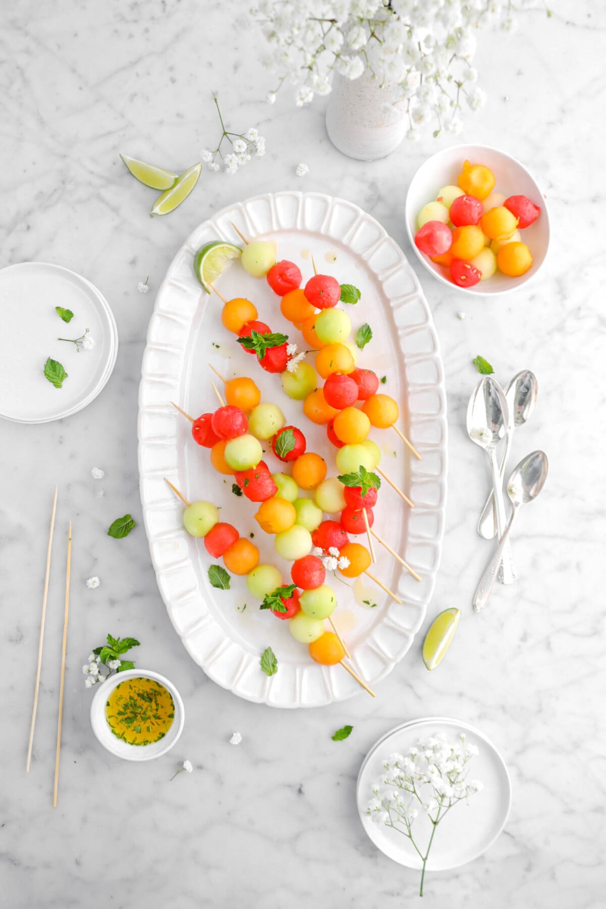 overhead shot of melon skewers on oval white platter with bowl of balled melons beside, spoons, and plates beside