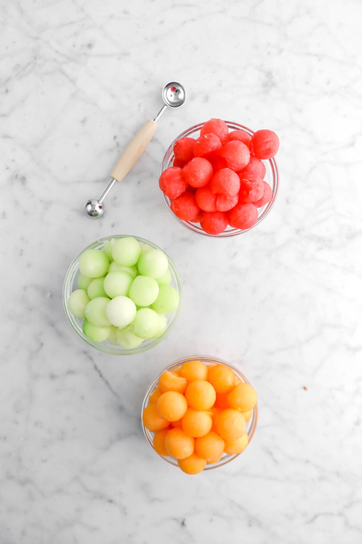 watermelon balls, honeydew balls, and cantaloupe balls in glass bowls on marble surface with melon baller beside