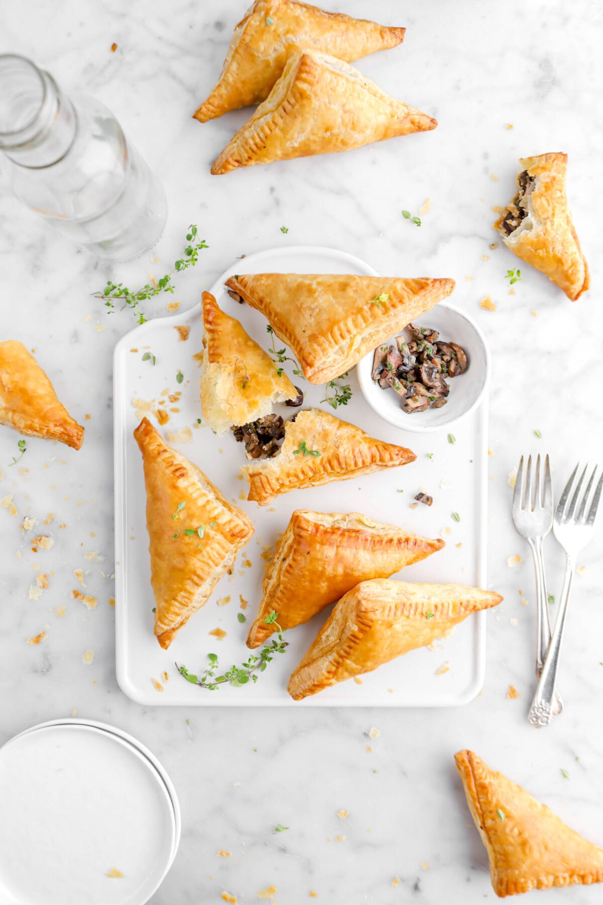 turnovers on white tray with bowl of mushrooms and thyme sprigs, more turnovers around on marble surface, with plates and forks beside
