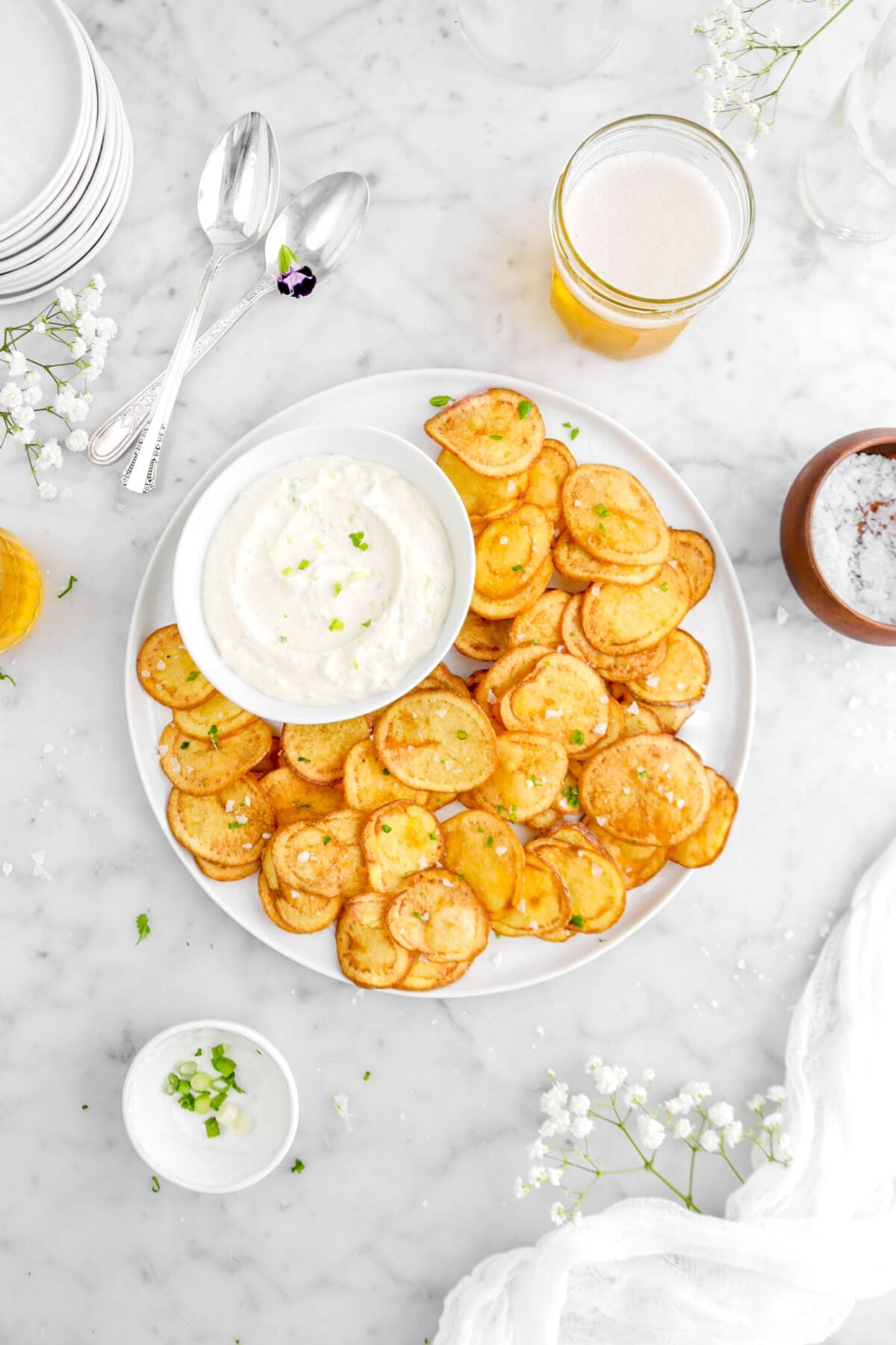 plate of potato chips with bowl of dip, glass of beer beside, wood bowl of salt, green onions around, and two spoons beside with purple flower