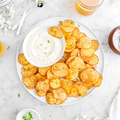 overhead shot of white plate full of potato chips with flakes salt and green onions on top, bowl of dip with potato chip in it, two spoons, and a glass of beer beside