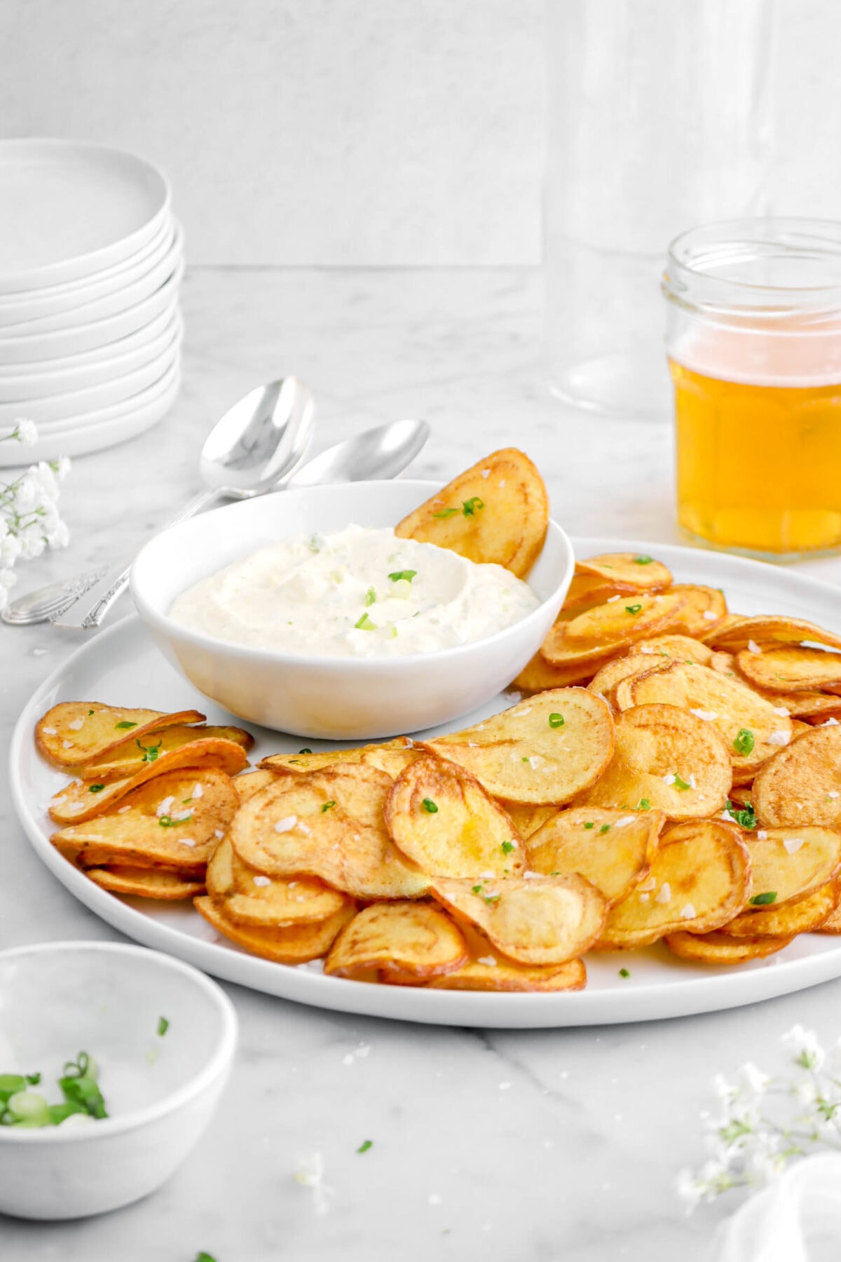 front shot of potaot chips on white plate with white bowl of dip and potato chip dipped in it, glass of jar with beer in it behind, stack of white plates, and bowl of chopped green onions