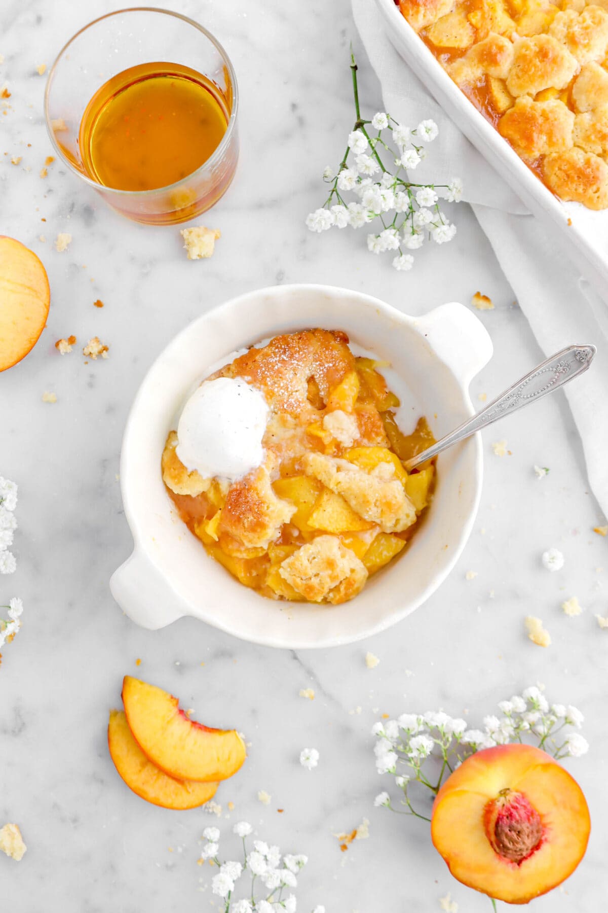peach cobbler in small ceramic pie pan with glass of bourbon and flowers around
