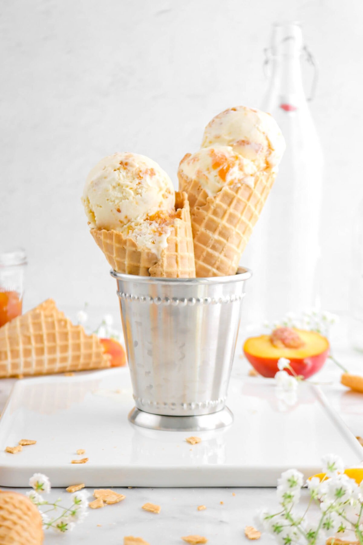 close up of two cones of peaches and cream ice cream in metal cup on square plate with flowers, waffle cones, and peaches around