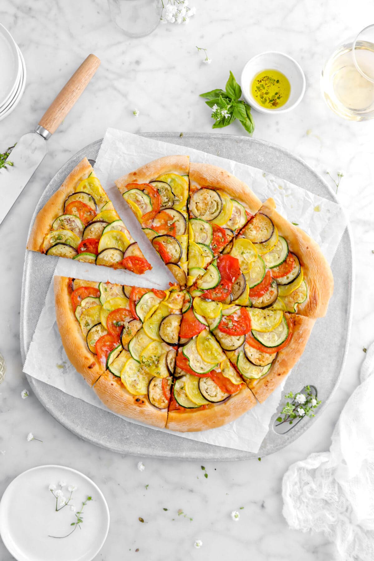 pizza on metal grill pan with top left corner piece pull away from pizza with fresh herbs, bowl of oil, and glass of wine beside