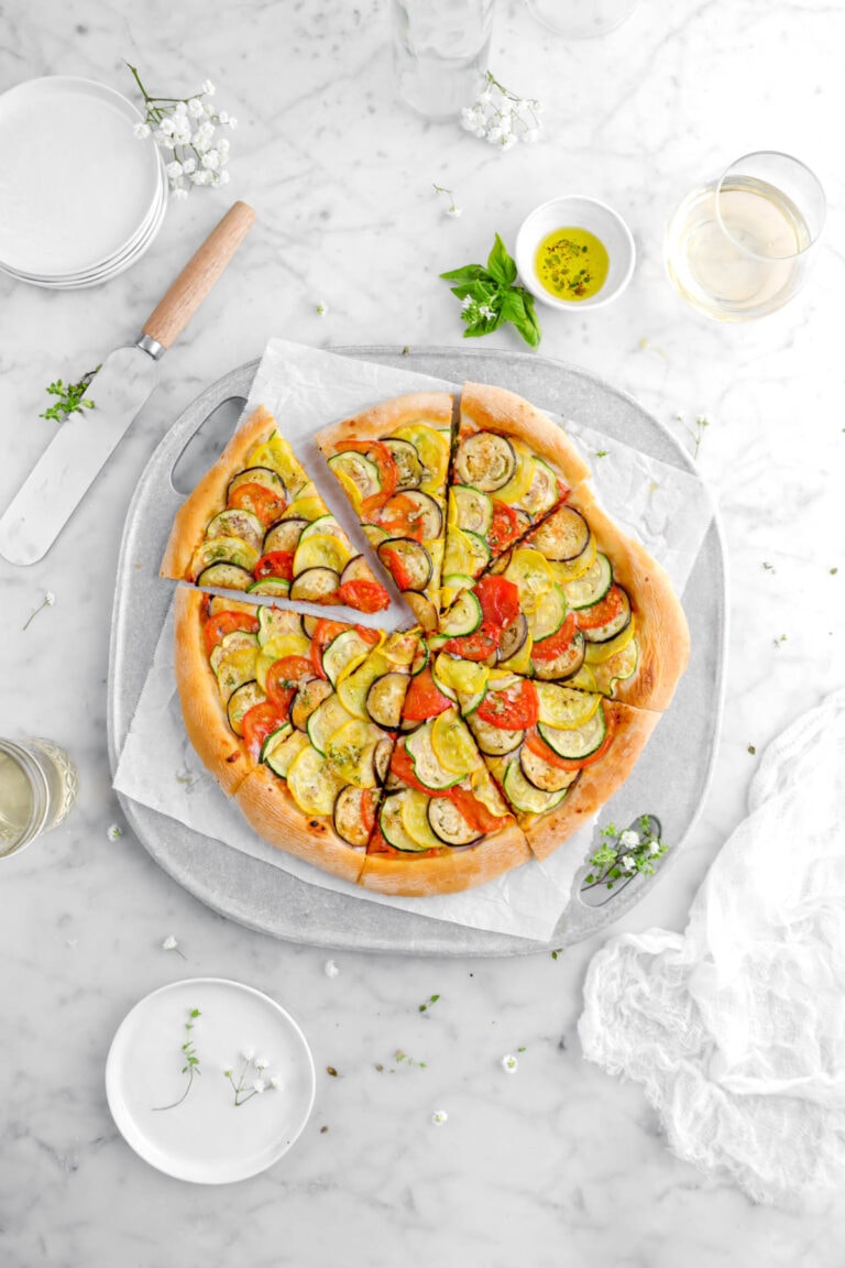 overhead shot of ratatouille pizza with top left corner slice pulled slightly away from rest of pizza on grill pan with fresh herbs, bowl of olive oil, two glasses of white wine, white plates, and pizza turner around on marble surface with white cheesecloth