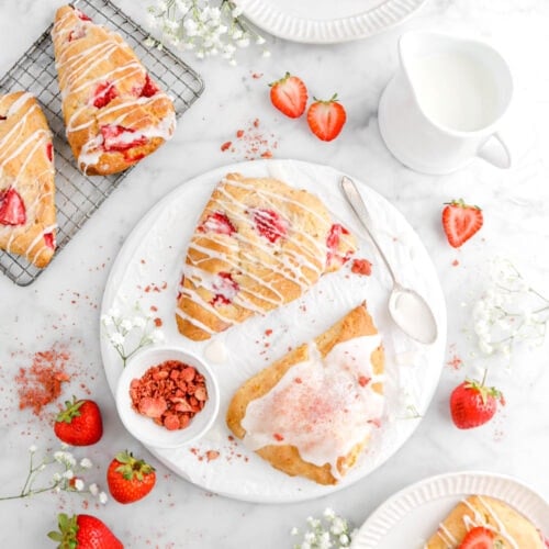 two scones on white plate with bowl of freeze dried strawberries beside and spoon, creamer, fresh strawberries, and more scones around
