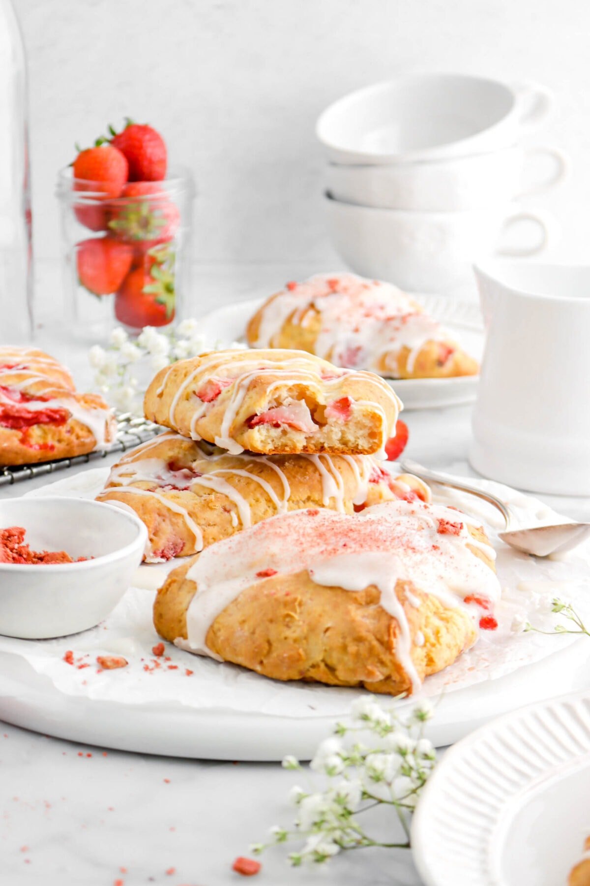 strawberry scone with bite missing stacked on another scone on upside downw hite palte with more scones around, jar of fresh strawberries, and stack of white coffee mugs